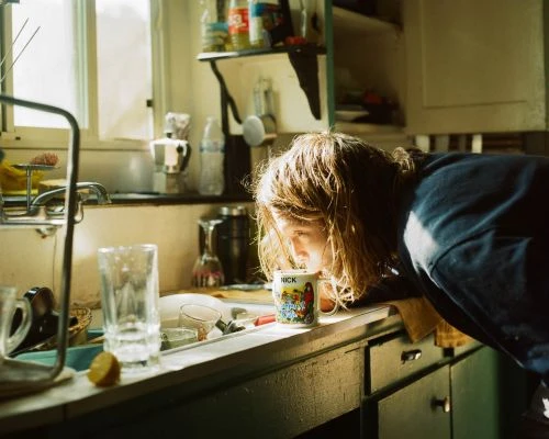 A person with long hair leans over a kitchen sink, drinking from a patterned mug in warm natural light.