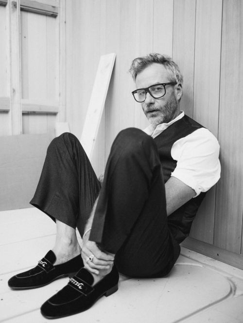 Black and white portrait of a man wearing glasses, a vest, and loafers, sitting on the floor with a neutral facial expression.