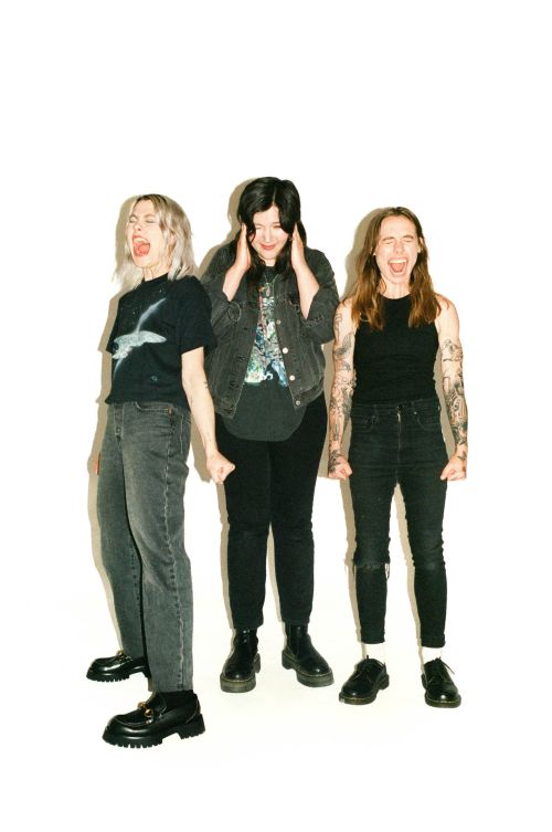 Three women stand indoors against a white background. The women wear casual dark clothing and boots. One woman is covering her ears.