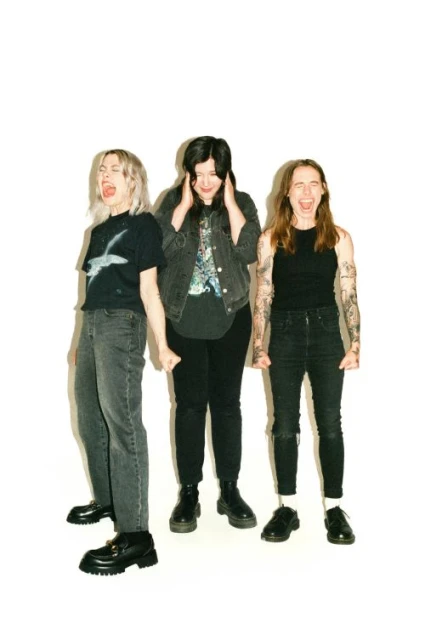 Three women stand indoors against a white background. The women wear casual dark clothing and boots. One woman is covering her ears.