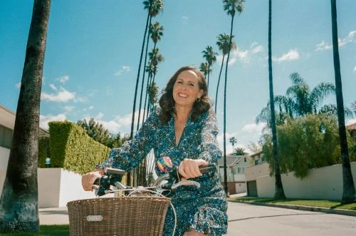 A woman in a blue patterned dress smiles while riding her bicycle with a basket on a tree-lined street on a sunny day.