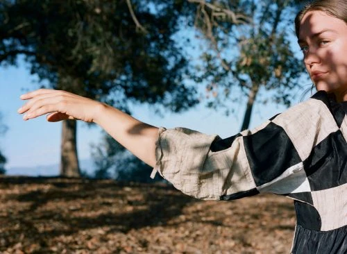 Adult female poses in a checker-patterned top, arm extended, amidst trees and light shadows in an outdoor setting during daytime.