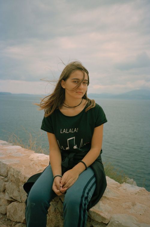 A young woman with long brown hair sits on a stone wall overlooking a body of water and distant mountains under a cloudy sky.