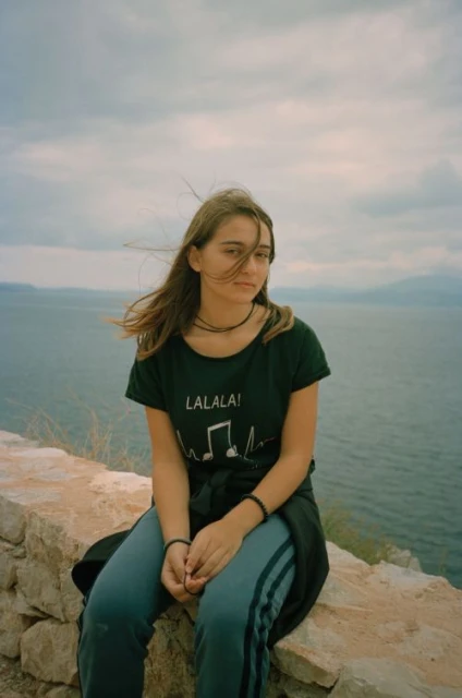 A young woman with long brown hair sits on a stone wall overlooking a body of water and distant mountains under a cloudy sky.