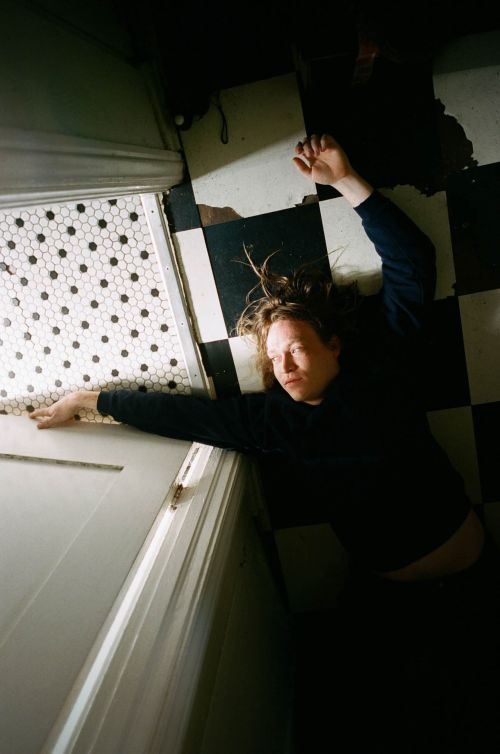 Adult with long brown hair lies on a black-and-white checkerboard tile floor next to a white door with hexagonal-tiled trim.