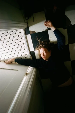 Adult with long brown hair lies on a black-and-white checkerboard tile floor next to a white door with hexagonal-tiled trim.