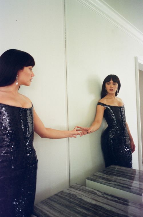 Woman with dark hair wearing a black sequin dress touching her reflection in a large wall mirror.