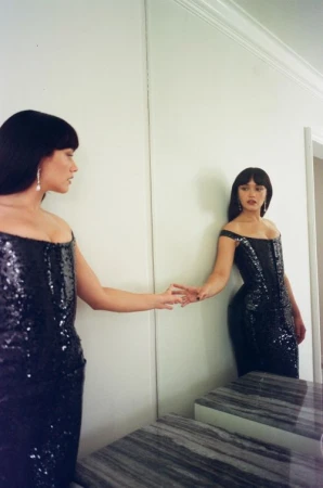 Woman with dark hair wearing a black sequin dress touching her reflection in a large wall mirror.
