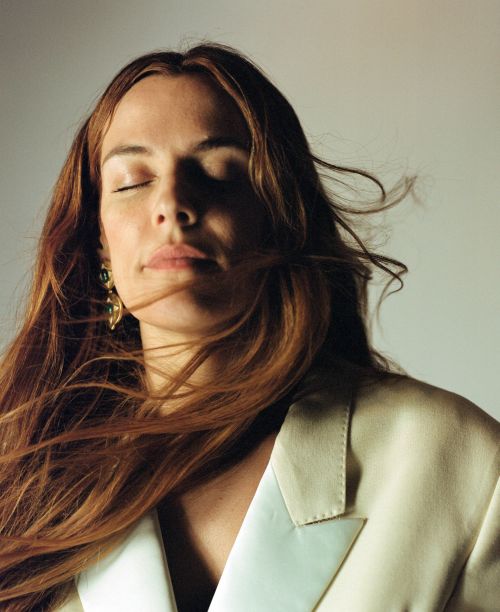 Portrait of a woman with auburn hair, closed eyes, and wearing a white blazer against a neutral background in soft light.