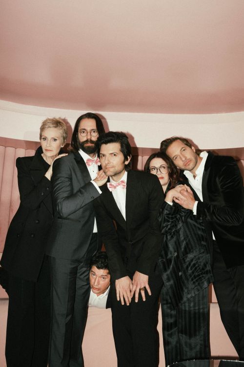 Six people in formal wear pose indoors against a pink backdrop; the group includes adult men and women with various poses and expressions.