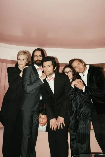Six people in formal wear pose indoors against a pink backdrop; the group includes adult men and women with various poses and expressions.