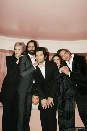 Six people in formal wear pose indoors against a pink backdrop; the group includes adult men and women with various poses and expressions.