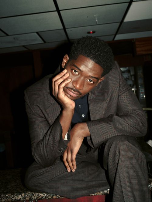An adult male in a suit sits on a countertop, resting his face in his hand with a thoughtful expression, set against a dark background.
