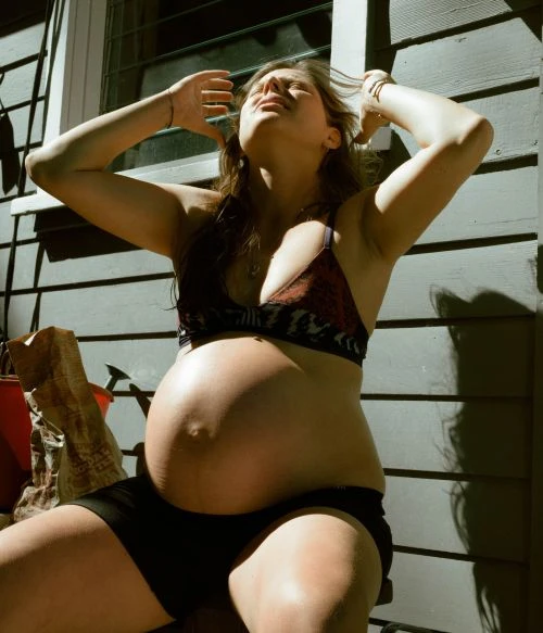 Pregnant woman sits outside in the sun wearing a bikini top and shorts. She has her arms raised above her head.