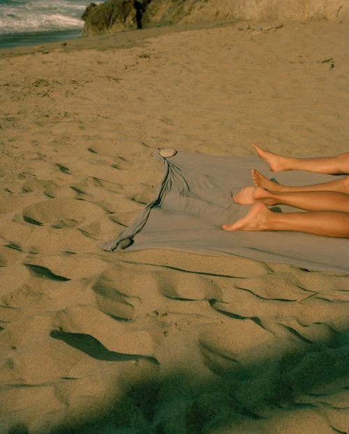 Two sets of legs lying on a blue blanket on a beach near the ocean under bright sunlight, outdoor setting, lifestyle photography.