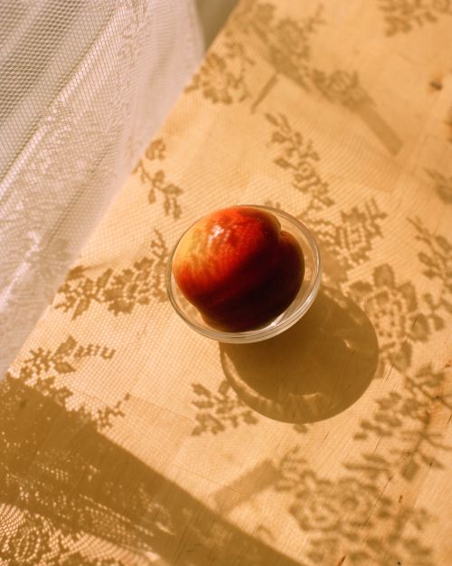 Close-up of a ripe peach in a glass bowl sitting on a beige floral-patterned tablecloth with lace curtains casting shadows.