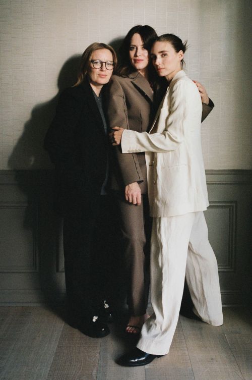Three adult women pose indoors wearing business suits; two embrace closely.  One woman wears eyeglasses. Neutral colors and studio lighting.