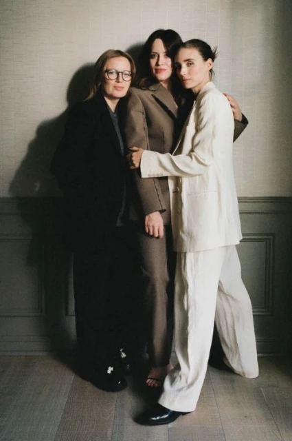 Three adult women pose indoors wearing business suits; two embrace closely.  One woman wears eyeglasses. Neutral colors and studio lighting.