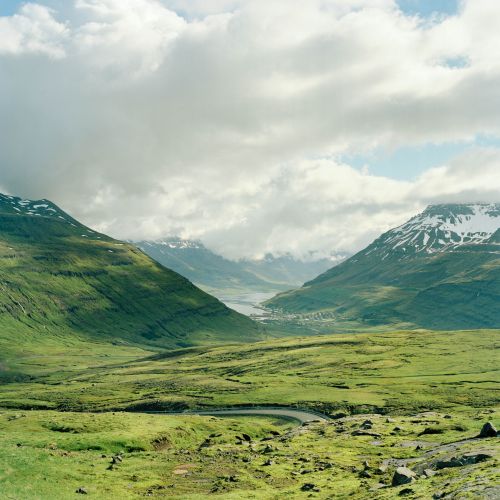 Scenic vista of lush green valley with winding road, snow-capped mountains, and cloudy sky, creating a peaceful landscape.