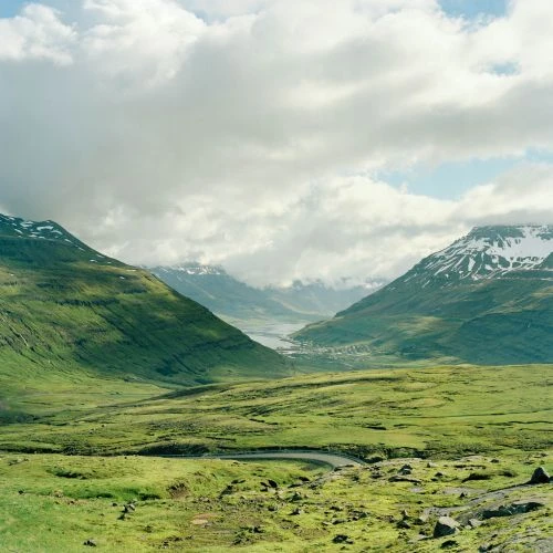 Scenic vista of lush green valley with winding road, snow-capped mountains, and cloudy sky, creating a peaceful landscape.
