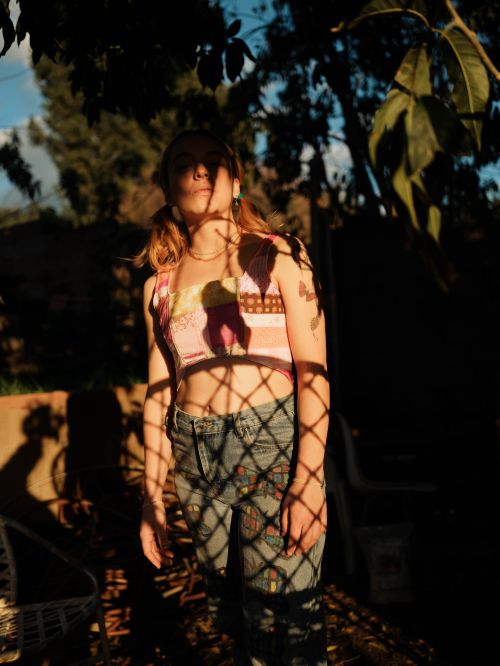 Young woman in a colorful top and patterned jeans standing in dappled sunlight with trees and foliage visible in the background.