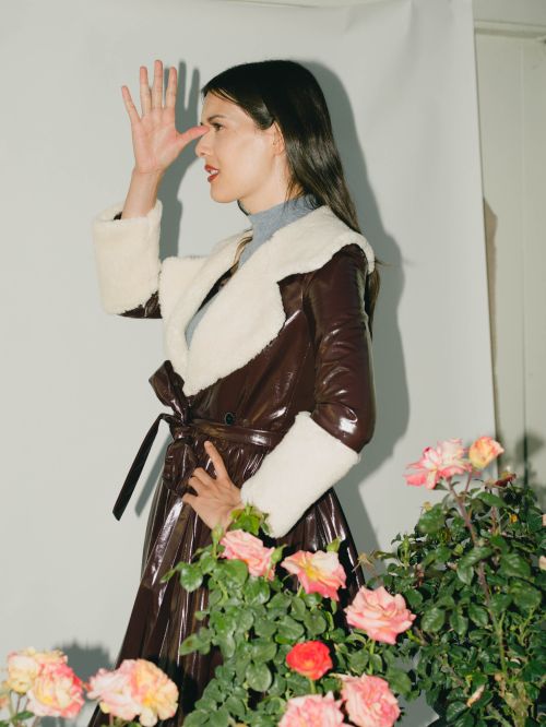 Woman in brown glossy belted coat with white fur collar posing behind pink roses, photographed by Chantal Anderson.