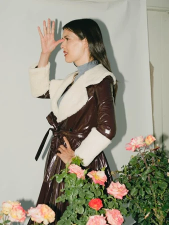 Woman in brown glossy belted coat with white fur collar posing behind pink roses, photographed by Chantal Anderson.