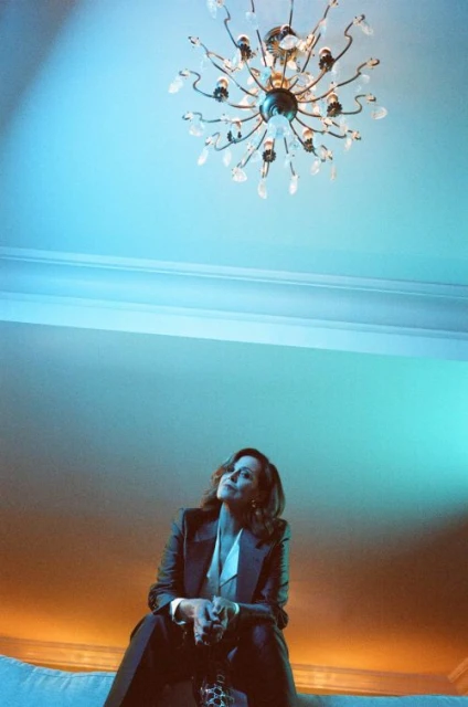A portrait shot of an adult woman with long brown hair, wearing a tan coat, posing against a blurred background of natural outdoor light.