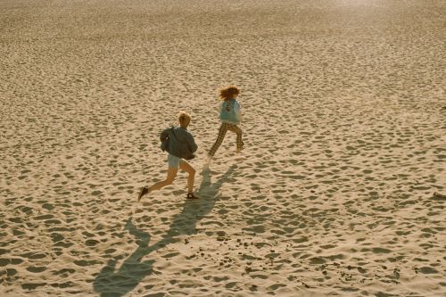 Two people running across a wide sandy beach under warm sunlight, photographed from an elevated high angle.