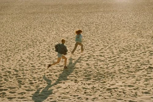 Two people running across a wide sandy beach under warm sunlight, photographed from an elevated high angle.