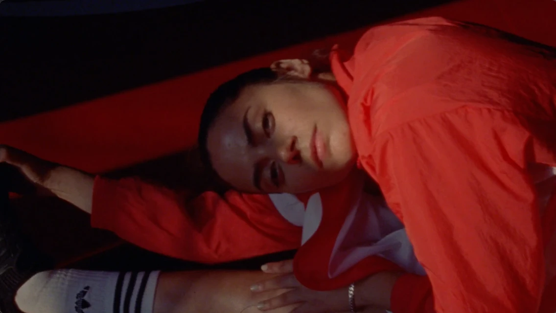A young adult woman in a red sports jacket lies on a red surface, photographed from a high angle in a dimly lit indoor setting.