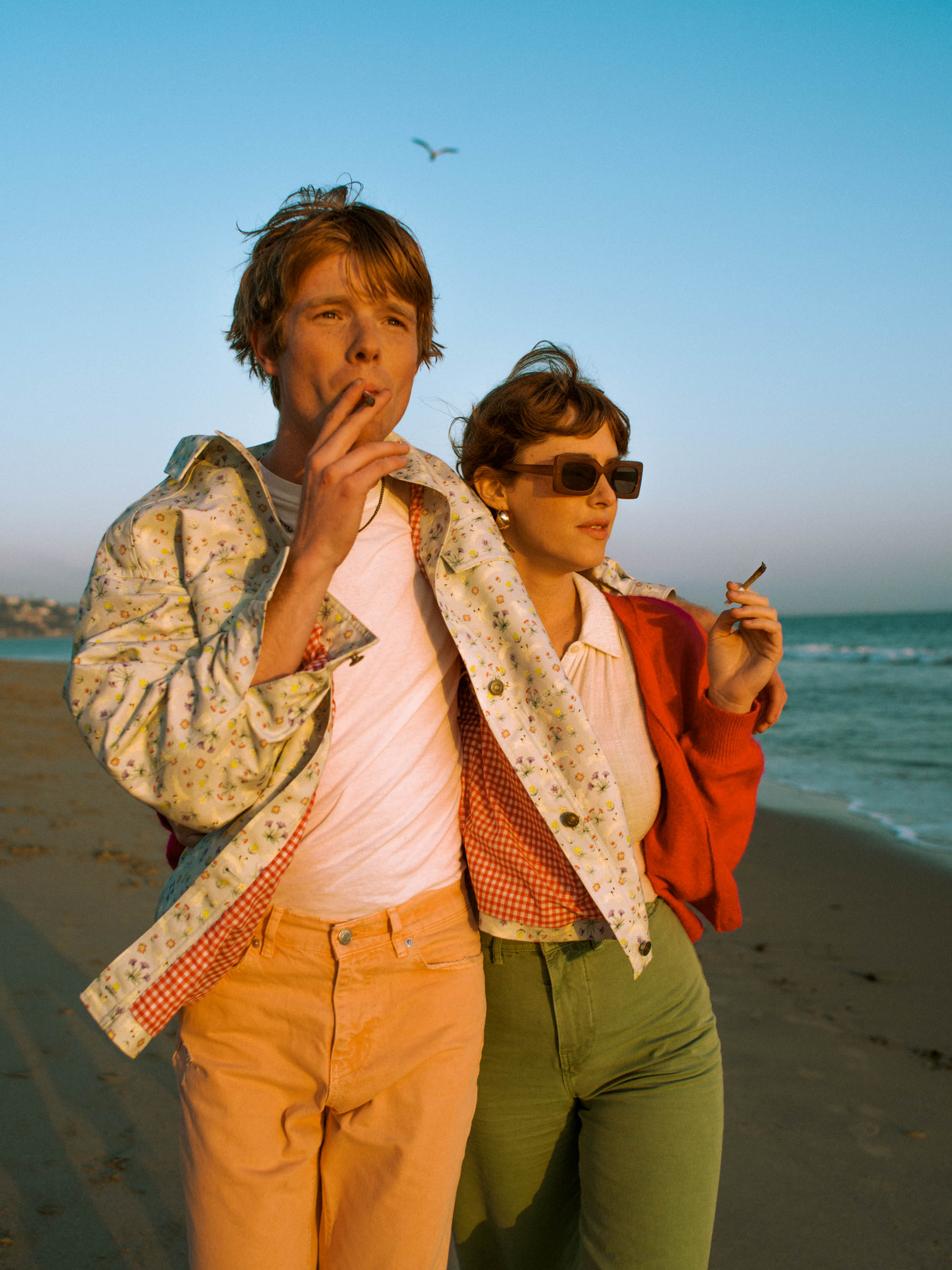 Two adults, a male and female, stand on a beach with ocean view. The man is smoking and wears a floral jacket. The woman wears sunglasses.