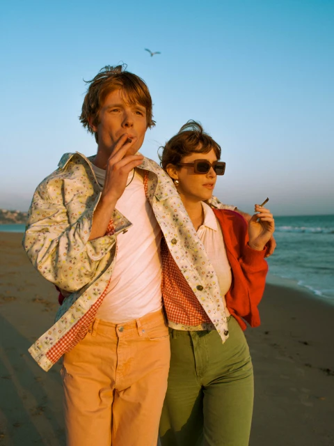Two adults, a male and female, stand on a beach with ocean view. The man is smoking and wears a floral jacket. The woman wears sunglasses.