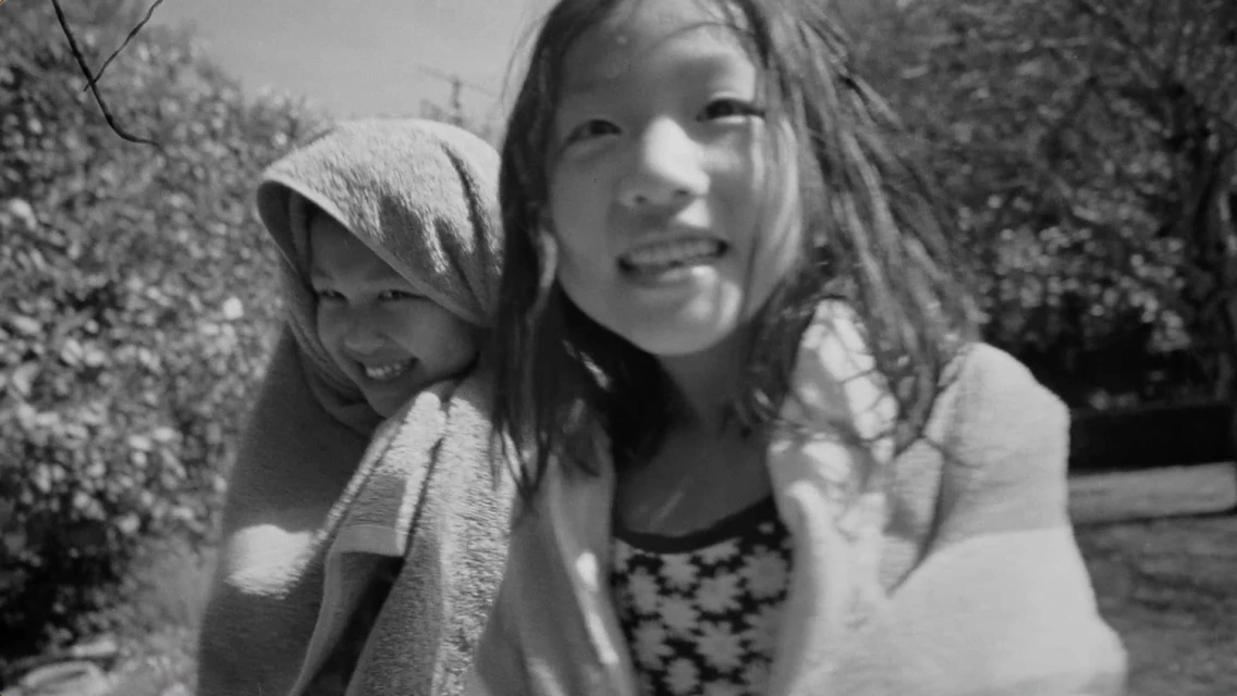 Black and white image of two smiling Asian girls, one wrapped in a towel, outdoors with blurred trees in the background.