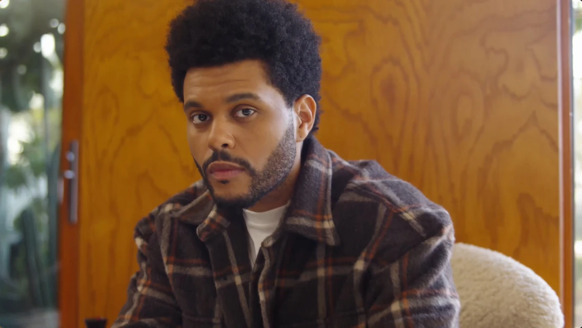 Portrait of an adult male with dark curly hair and a plaid shirt, set against a blurred interior with wood paneling. Head and shoulders shot.