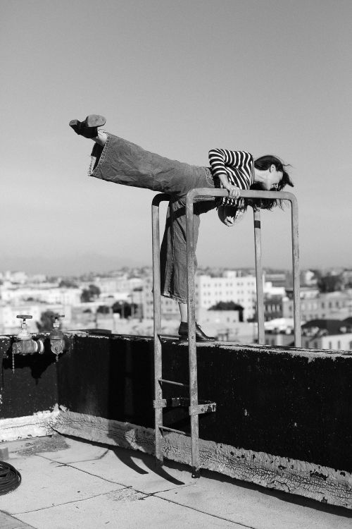 Black and white photo of a woman in striped shirt and wide-leg pants balancing on a metal ladder on a rooftop with city views.