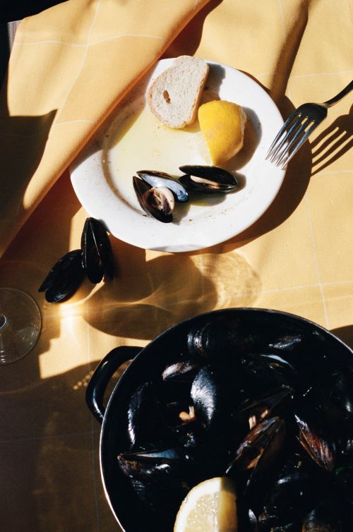Overhead shot of mussels served on a plate and in a pot, accompanied by a lemon wedge and slice of bread on a yellow tablecloth.