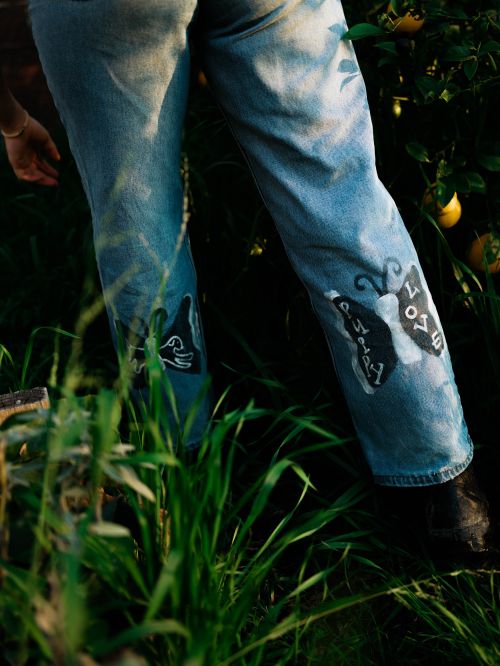 Close-up of custom-painted jeans featuring artistic designs and the word "Love," worn outdoors amidst lush greenery.