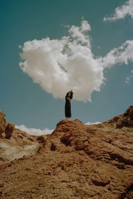 Adult woman in a patterned dress stands on a red-rock hill beneath a large white cloud in a landscape scene against a teal sky.