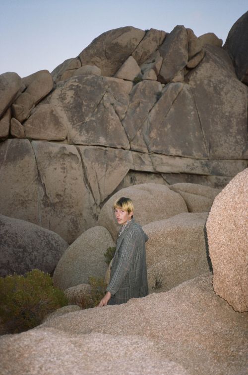 A person stands among large rocks in a desert landscape, wearing a grey jacket, creating a blend with the natural surroundings.