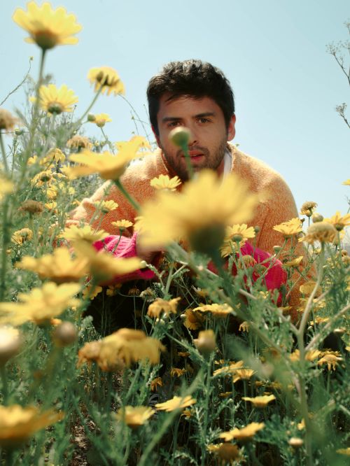 Adult male in a field of yellow daisy flowers, medium shot, low angle, wearing an orange sweater and pink gloves, against blue sky.