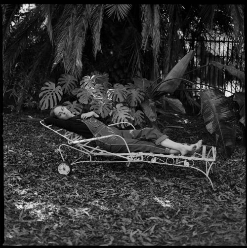 Woman in overalls lounges on a metal chaise lounge surrounded by leafy plants. Black and white medium shot with natural light.