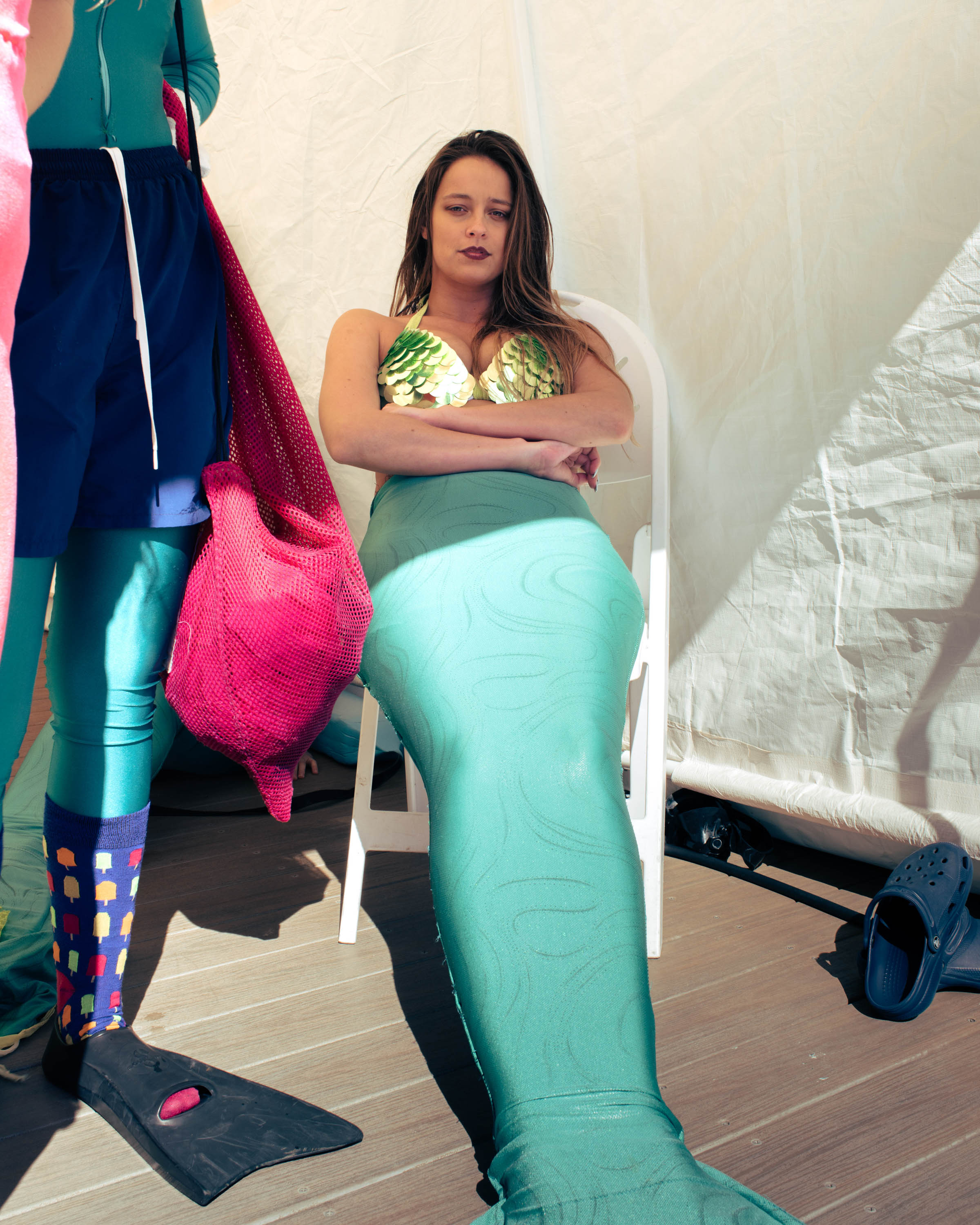 A woman with a green mermaid tail and a shell bra sits in a chair, crossing her arms, while standing next to people in fins.