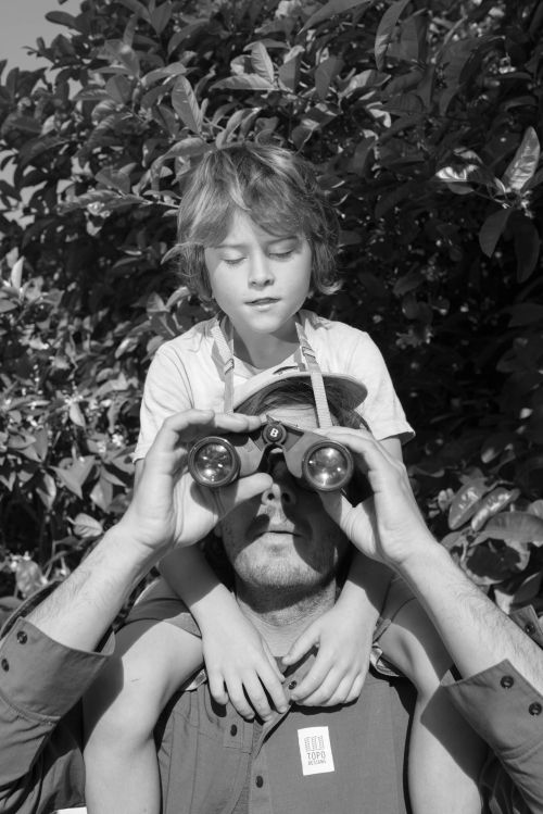 A child sits on an adult's shoulders, both looking through binoculars in front of a leafy background in this black and white outdoor shot.