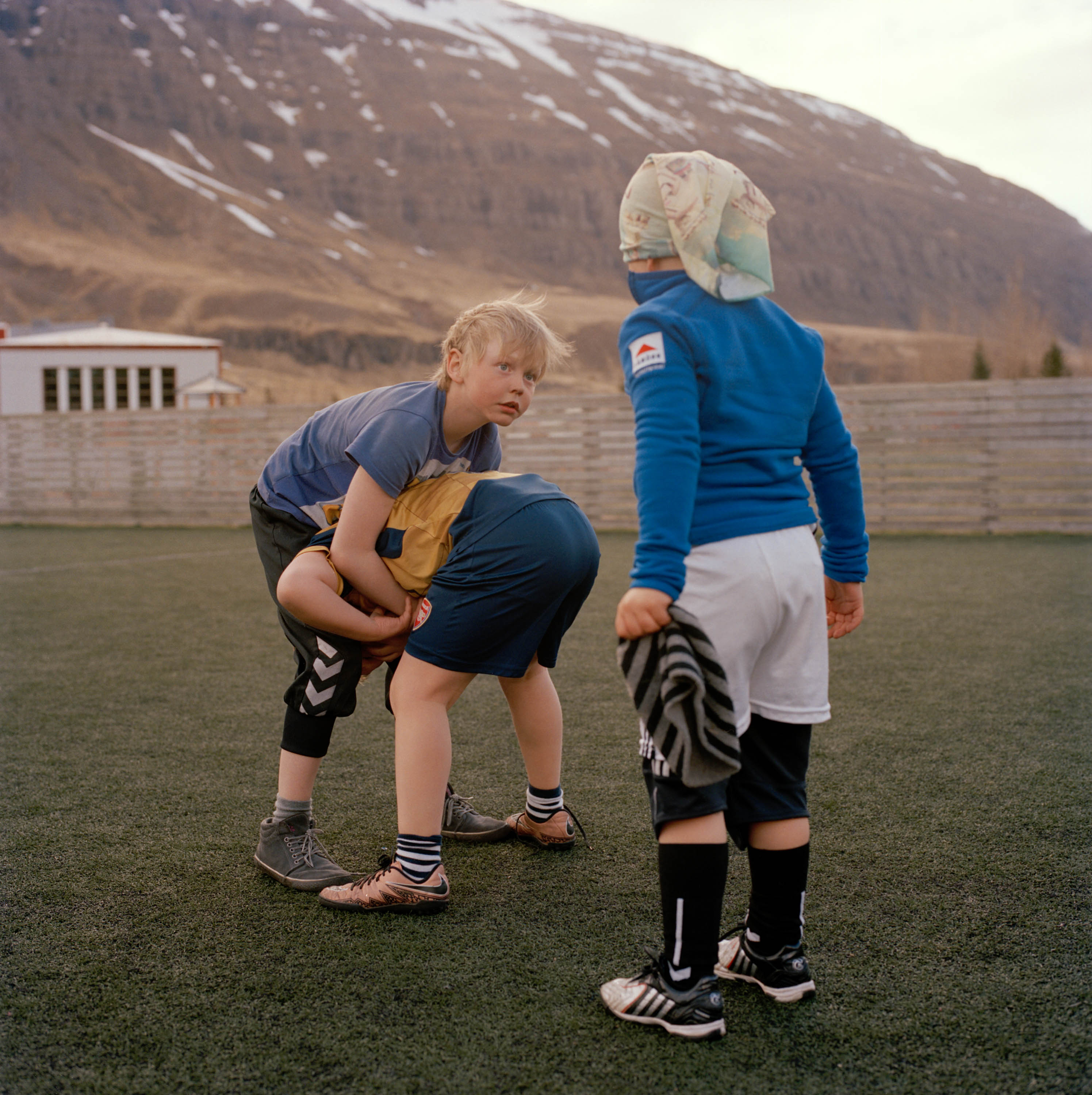 Three kids playing a game on a soccer field with mountains in the background. One boy has another on his back, facing a third boy.