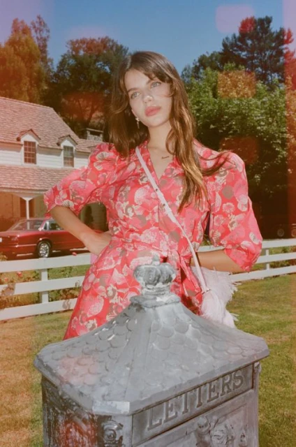Young woman in a floral dress stands outdoors next to a vintage mailbox, with a car and house visible in the background.