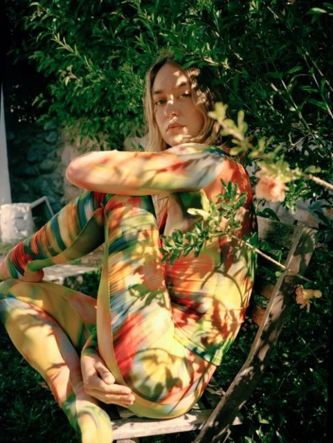 Adult woman with long blonde hair sitting cross-legged on a wooden chair in a garden, wearing a colorful tie-dye bodysuit.