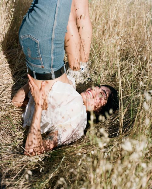 Woman in white top and blue jeans lying in a field of tall grass with another person standing over her outdoors on a sunny day.