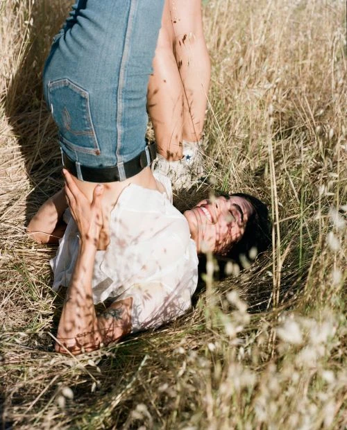 Woman in white top and blue jeans lying in a field of tall grass with another person standing over her outdoors on a sunny day.