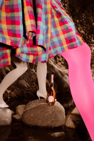 Legs of a person wearing bright pink tights and another in white stockings with heels near a bottle of wine on a rock